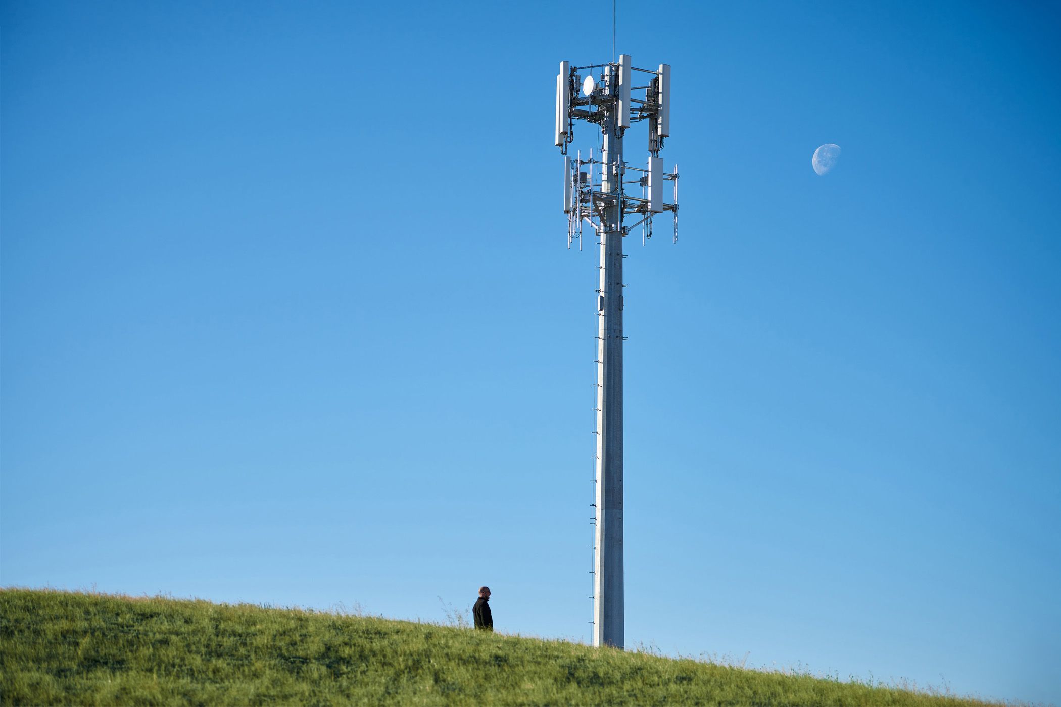 5G mmWave: A tecnologia mais rápida que vai mudar sua conexão 🚀⚡ 9 Torre de celular em uma colina com uma pessoa em pé abaixo e uma lua diurna no céu.