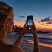 Fotografando a Lua com o iPhone: dicas para capturar detalhes 30 Fotos da lua com iPhone - Mulher fotografando a lua cheia com um iPhone em uma praia ao pôr do sol, usando truques para capturar seus detalhes.